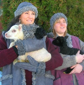 Två kvinnor iklädda västar, vantar och mössor av vackert, lockiga grå fårskinn. I famnen håller de varsitt lamm.